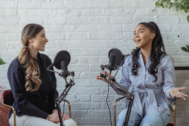 Două femei stau și discută în fața a două microfoane cu filtre pop.
